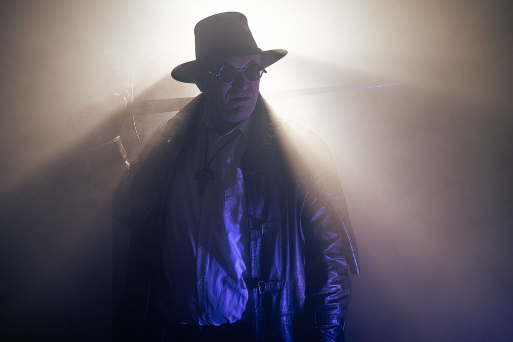 Van Helsing in a hat and leather coat with a sword, backlit through heavy fog